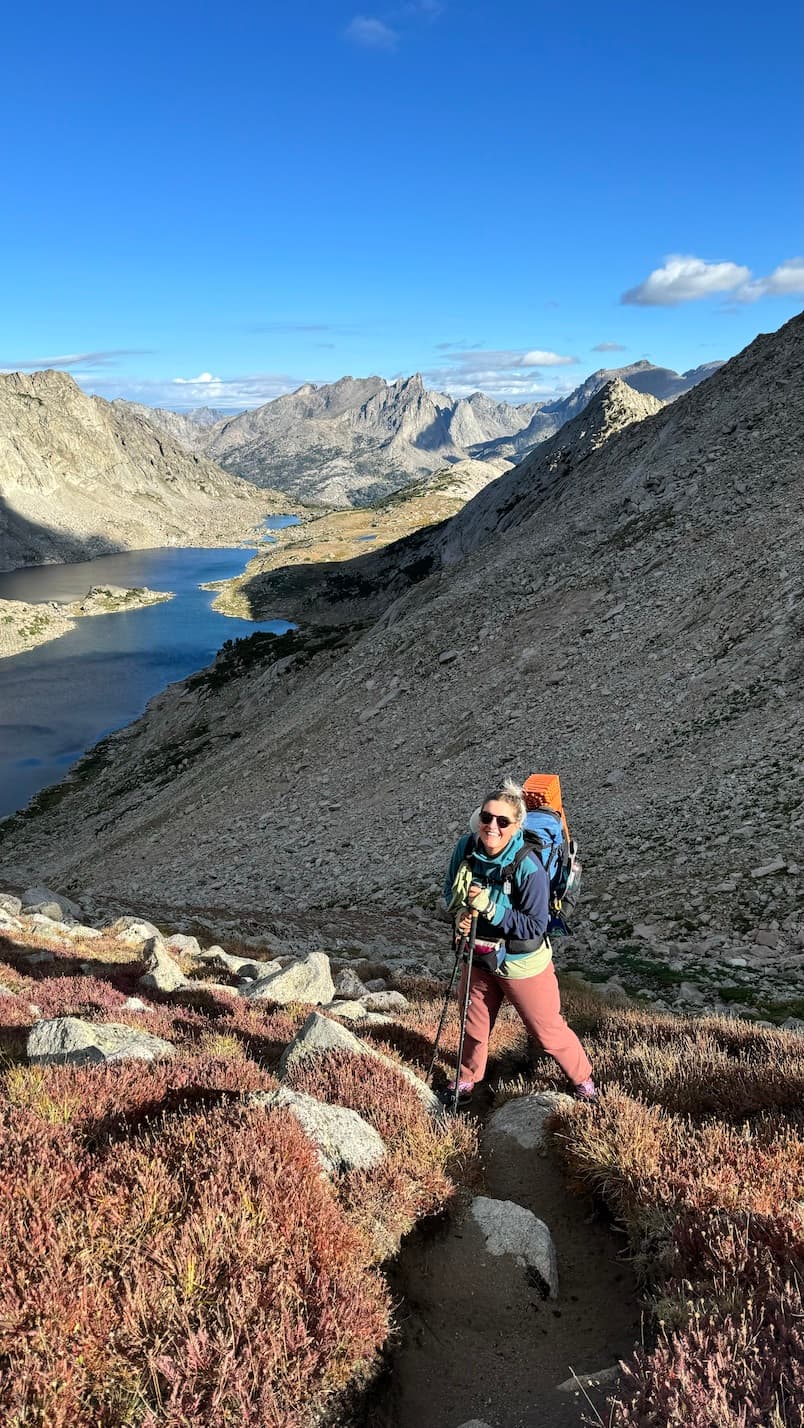 Photo of Katy nearing the top of a mountain pass in the Wind River Range near sunrise, many mountains and lakes behind her